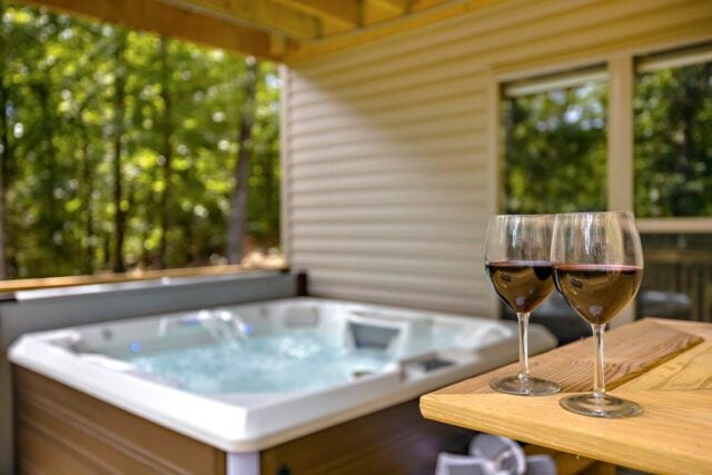 Relaxing outdoor hot tub with two glasses of wine on a wooden table, surrounded by lush green trees.