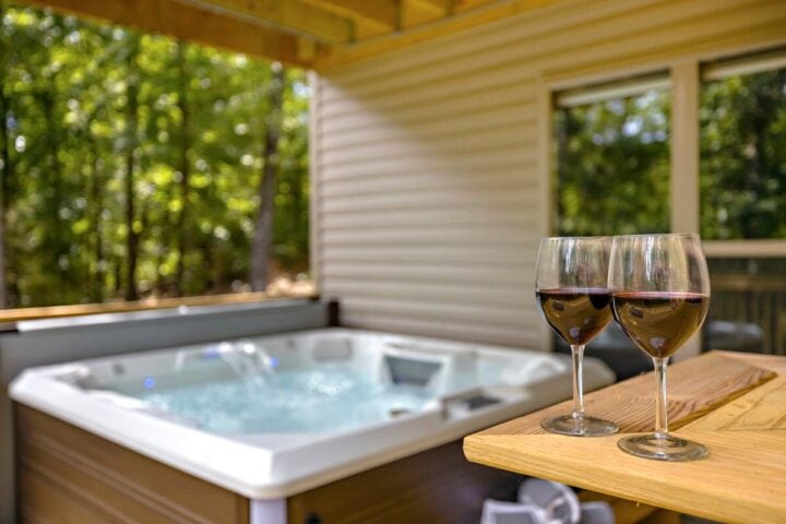 Relaxing outdoor hot tub with two glasses of wine on a wooden table, surrounded by lush green trees.