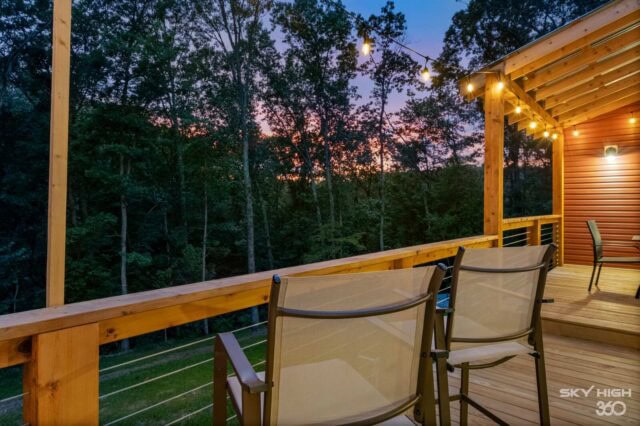 Cozy cabin balcony overlooking wooded landscape at sunset with outdoor seating and string lights.